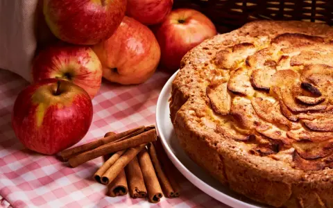 Torta mele, noci e cannella dorata, con mele fresche e bastoncini di cannella su una tovaglia a quadri rosa.