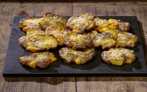 Patate schiacciate al forno, dorate e croccanti, cosparse di formaggio grattugiato e spezie, servite su una pietra scura.