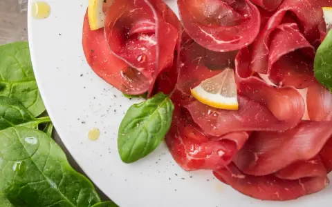 Fette sottili di bresaola di manzo su un piatto bianco, guarnite con spinaci freschi, spicchi di limone e un filo d'olio.