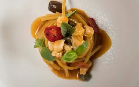Un piatto di pasta con il pesce, condita con pomodorini, olive, capperi e basilico, servita su un piatto bianco.