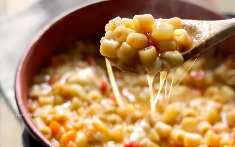 Un cucchiaio solleva una porzione fumante di pasta e patate veloce, filante di formaggio fuso, in una ciotola di terracotta.