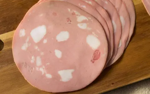 Fette di mortadella su tagliere di legno. Scopri come si fa la mortadella, un salume rosa con cubetti di grasso bianco.