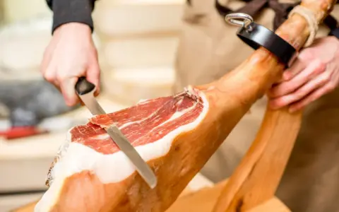 Mani esperte dimostrano come tagliare il prosciutto crudo con un coltello affilato, creando fette sottili e gustose.