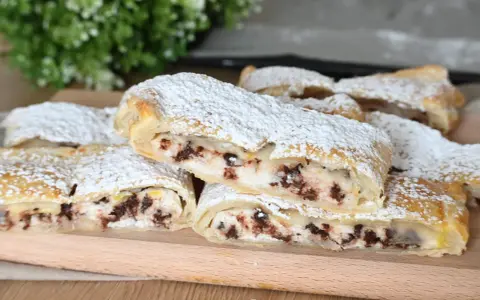 Rotolo di sfoglia con ricotta dolce e gocce di cioccolato, spolverato di zucchero a velo, su un tagliere di legno.