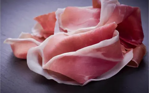 Fette di prosciutto crudo, un insaccato dal colore rosato e venature bianche, disposte su una superficie scura.