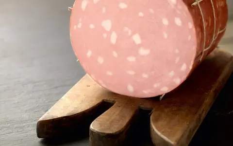 Una fetta di mortadella è cotta, con i suoi tipici cubetti bianchi di grasso, posata su un tagliere di legno.