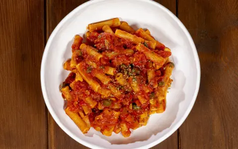 Un piatto di pasta alla pizzaiola, con rigatoni conditi con sugo di pomodoro, capperi e origano, servito in una ciotola bianca su un tavolo di legno scuro.