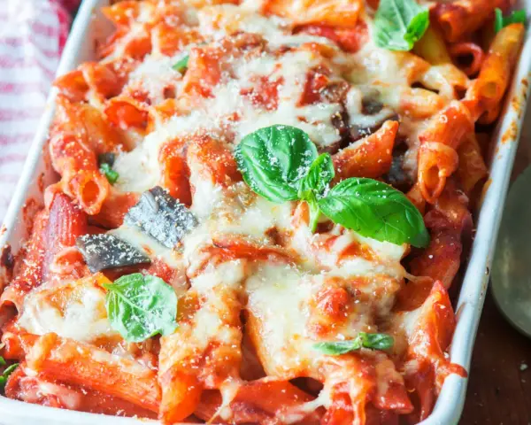 Piatto fumante di pasta al forno con melanzane, pomodoro e abbondante formaggio fuso, guarnito con foglie di basilico fresco.