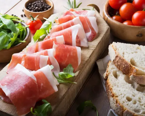 Fette di prosciutto crudo su tagliere con rucola, pomodorini e pane. Scopri come utilizzare il grasso del prosciutto crudo per insaporire i tuoi piatti.