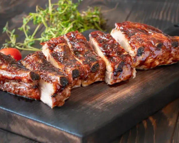 Rosticciana al forno morbida, succulenta e glassata, servita su tagliere scuro con pomodorini e rosmarino.