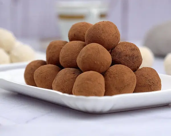 Montagna di tartufi al cioccolato senza panna, spolverati di cacao, su un piatto bianco. Sullo sfondo, altri dolcetti chiari.
