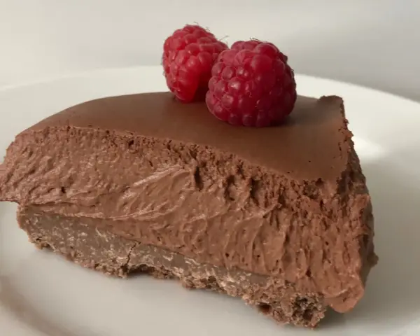 Una fetta di torta mousse al cioccolato, soffice e golosa, guarnita con due lamponi freschi su un piatto bianco.