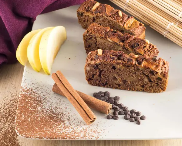 Fette di plumcake mela e cannella su piatto bianco, con mele, cannella, gocce di cioccolato e cacao in polvere.