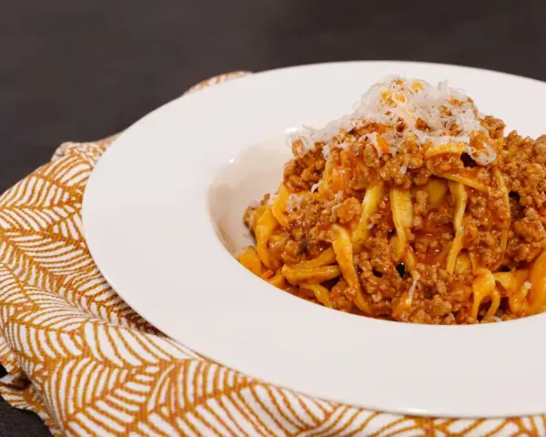 Tagliatelle ricche con sugo finto e parmigiano grattugiato, servite su un piatto bianco con tovagliolo decorato.