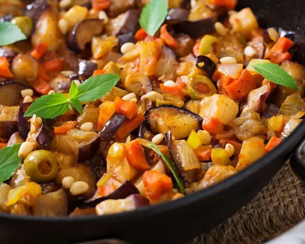 Caponata ricetta: melanzane, carote, olive e pinoli in padella, guarnita con basilico fresco. Un piatto colorato e saporito.