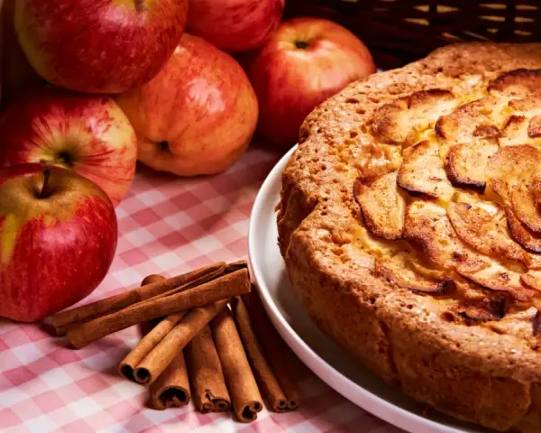 Torta mele, noci e cannella dorata, con mele fresche e bastoncini di cannella su una tovaglia a quadri rosa.