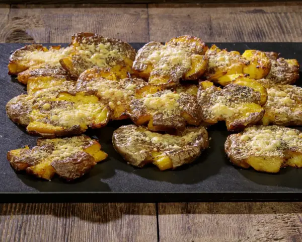 Patate schiacciate al forno, dorate e croccanti, cosparse di formaggio grattugiato e spezie, servite su una pietra scura.
