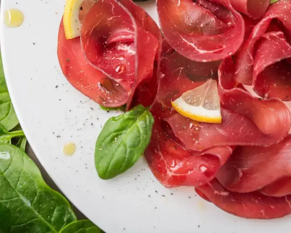 Fette sottili di bresaola di manzo su un piatto bianco, guarnite con spinaci freschi, spicchi di limone e un filo d'olio.