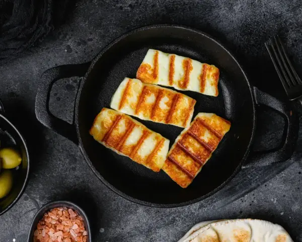 Quattro fette di galbanino alla piastra dorate e striate, servite in una padella di ghisa, con olive e sale rosa a lato.