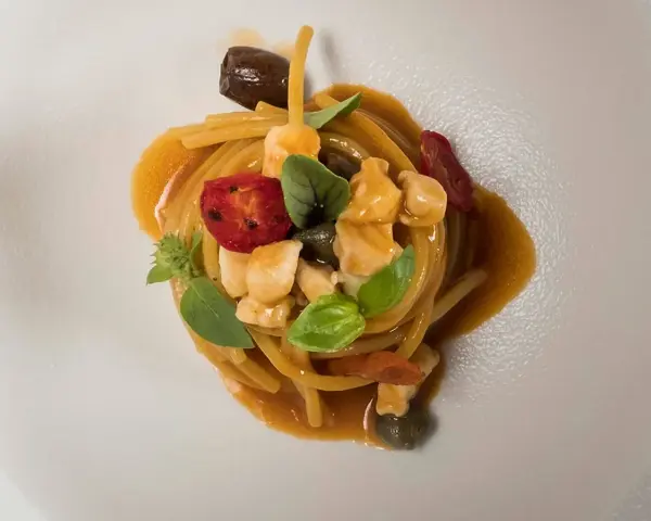 Un piatto di pasta con il pesce, condita con pomodorini, olive, capperi e basilico, servita su un piatto bianco.