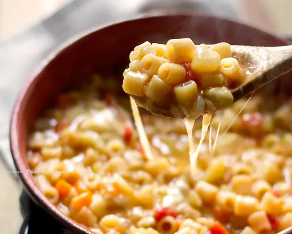 Un cucchiaio solleva una porzione fumante di pasta e patate veloce, filante di formaggio fuso, in una ciotola di terracotta.