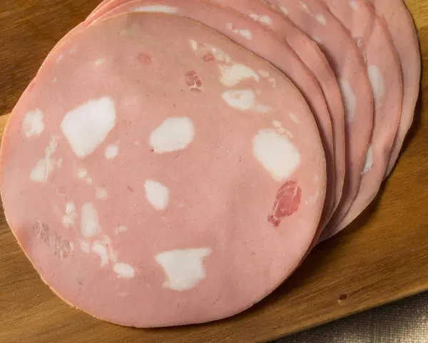 Fette di mortadella su tagliere di legno. Scopri come si fa la mortadella, un salume rosa con cubetti di grasso bianco.