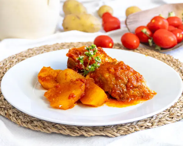 Piatto di cosciotto di tacchino in umido con patate, guarnito con erbe aromatiche. Sullo sfondo, pomodori e patate fresche.