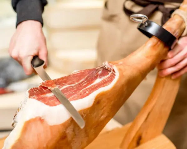 Mani esperte dimostrano come tagliare il prosciutto crudo con un coltello affilato, creando fette sottili e gustose.
