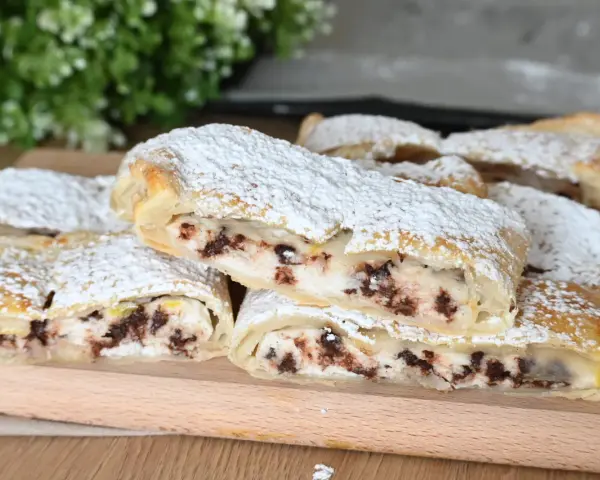 Rotolo di sfoglia con ricotta dolce e gocce di cioccolato, spolverato di zucchero a velo, su un tagliere di legno.