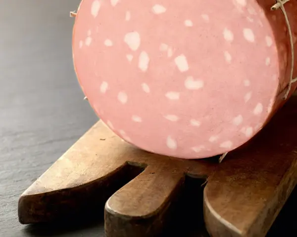 Una fetta di mortadella è cotta, con i suoi tipici cubetti bianchi di grasso, posata su un tagliere di legno.