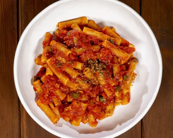 Un piatto di pasta alla pizzaiola, con rigatoni conditi con sugo di pomodoro, capperi e origano, servito in una ciotola bianca su un tavolo di legno scuro.