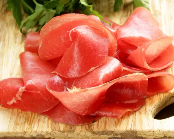 Fettine sottili di salume simile alla bresaola, rosse e lucide, disposte su un tagliere di legno con un ciuffo di prezzemolo.