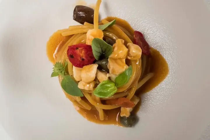 Un piatto di pasta con il pesce, condita con pomodorini, olive, capperi e basilico, servita su un piatto bianco.