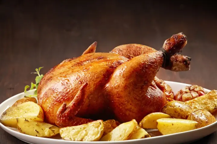 Un succulento pollo allo spiedo dorato, servito con patate arrosto e aglio su un piatto bianco.