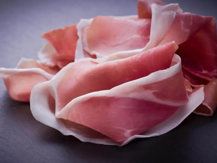 Fette di prosciutto crudo, un insaccato dal colore rosato e venature bianche, disposte su una superficie scura.