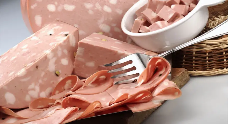 Fetta di mortadella con pistacchi e pepe, servita su un tagliere con fette arrotolate e cubetti in una ciotola.