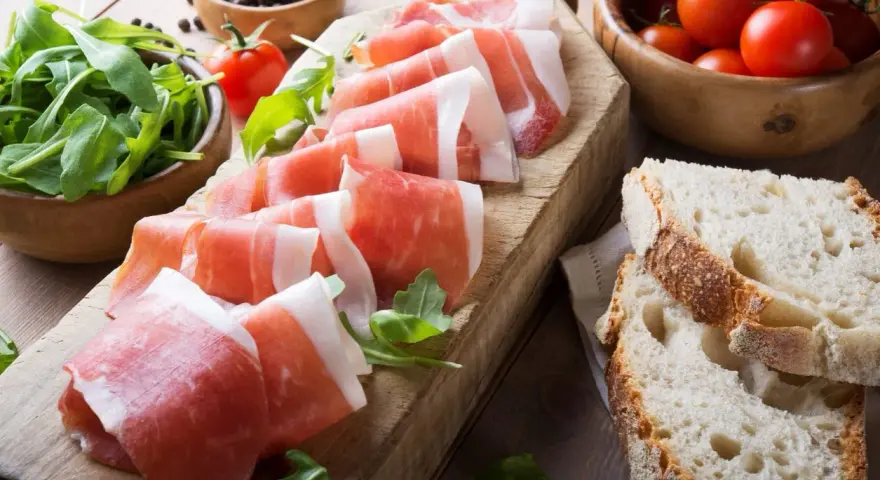 Fette di prosciutto crudo su tagliere con rucola, pomodorini e pane. Scopri come utilizzare il grasso del prosciutto crudo per insaporire i tuoi piatti.