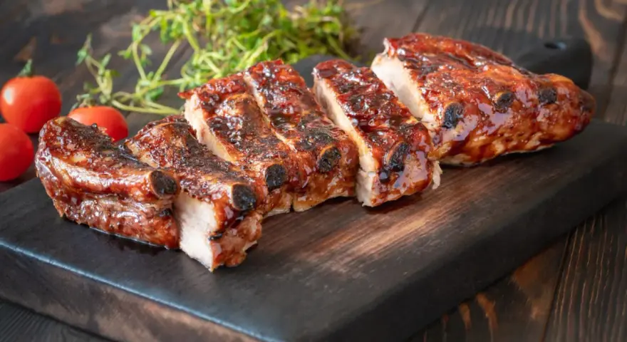 Rosticciana al forno morbida, succulenta e glassata, servita su tagliere scuro con pomodorini e rosmarino.