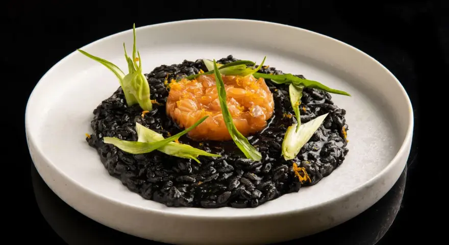 Un piatto di risotto al nero di seppia, guarnito con tartare di salmone e germogli verdi.