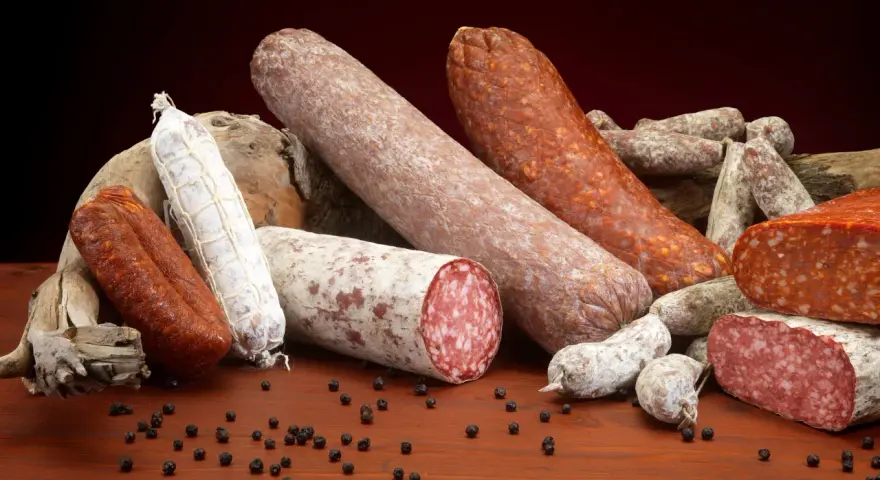 Un assortimento di salumi, tra cui spicca una grande fetta di carne insaccata, su un tavolo di legno con grani di pepe nero.