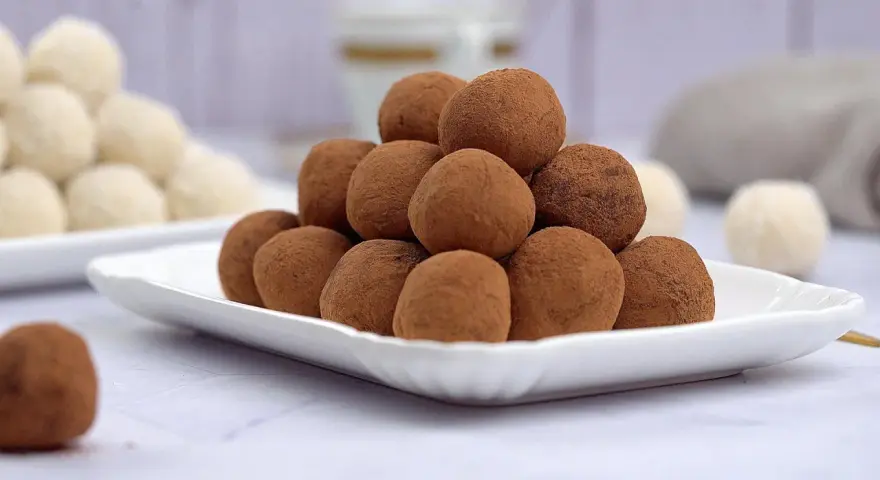 Montagna di tartufi al cioccolato senza panna, spolverati di cacao, su un piatto bianco. Sullo sfondo, altri dolcetti chiari.