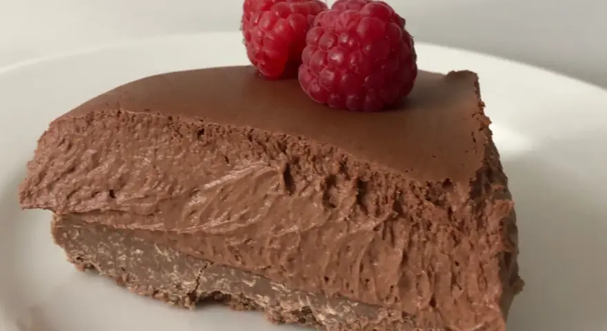 Una fetta di torta mousse al cioccolato, soffice e golosa, guarnita con due lamponi freschi su un piatto bianco.