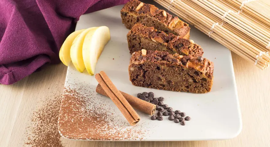 Fette di plumcake mela e cannella su piatto bianco, con mele, cannella, gocce di cioccolato e cacao in polvere.