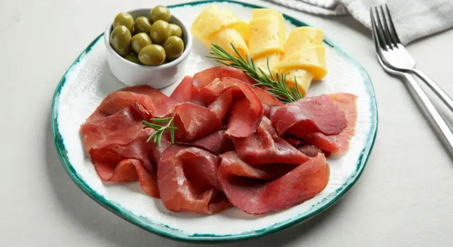 Bresaola con formaggio da abbinare, olive verdi e rosmarino su un piatto.