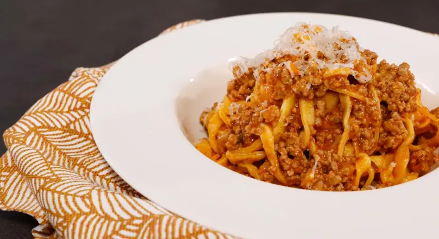 Tagliatelle ricche con sugo finto e parmigiano grattugiato, servite su un piatto bianco con tovagliolo decorato.