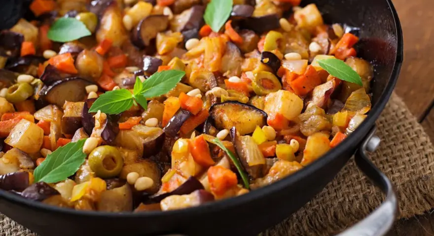 Caponata ricetta: melanzane, carote, olive e pinoli in padella, guarnita con basilico fresco. Un piatto colorato e saporito.