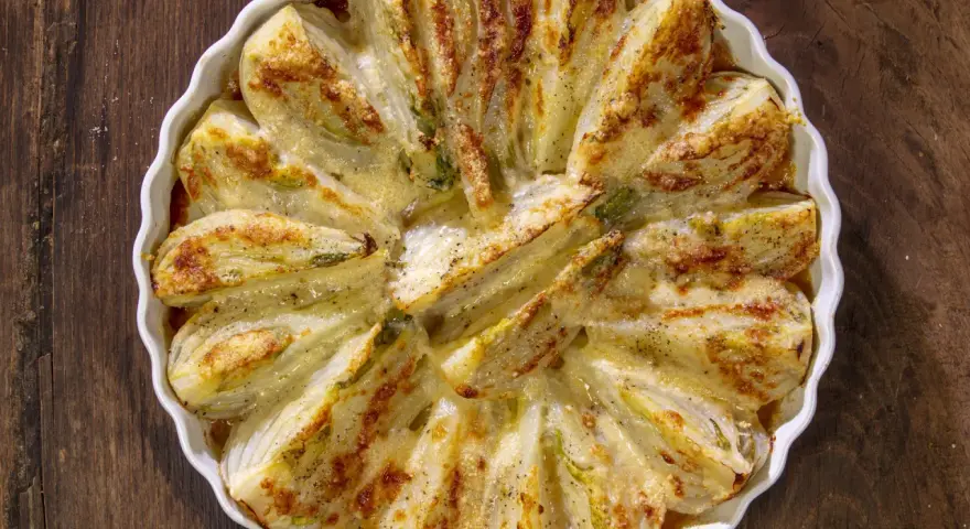 Finocchi al forno dorati e gratinati, serviti in una pirofila bianca su un tavolo di legno scuro.