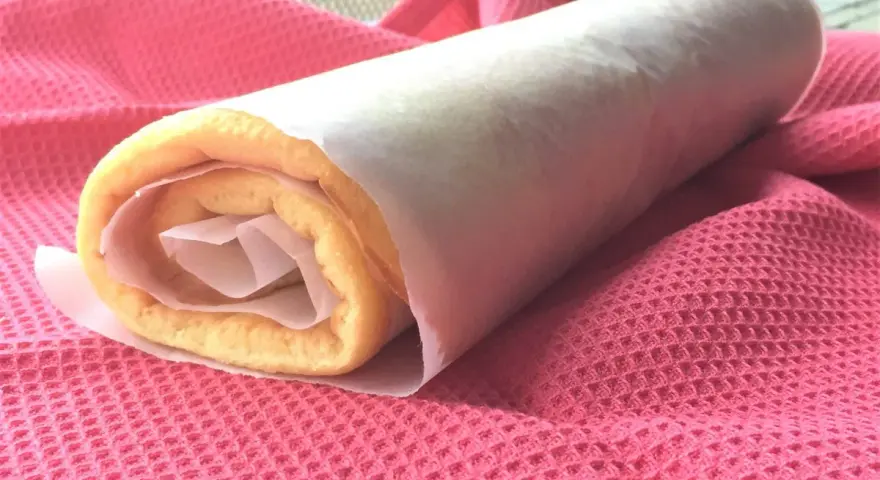 Rotolo di torta biscotto appena preparato, avvolto in carta forno, adagiato su un panno rosa.