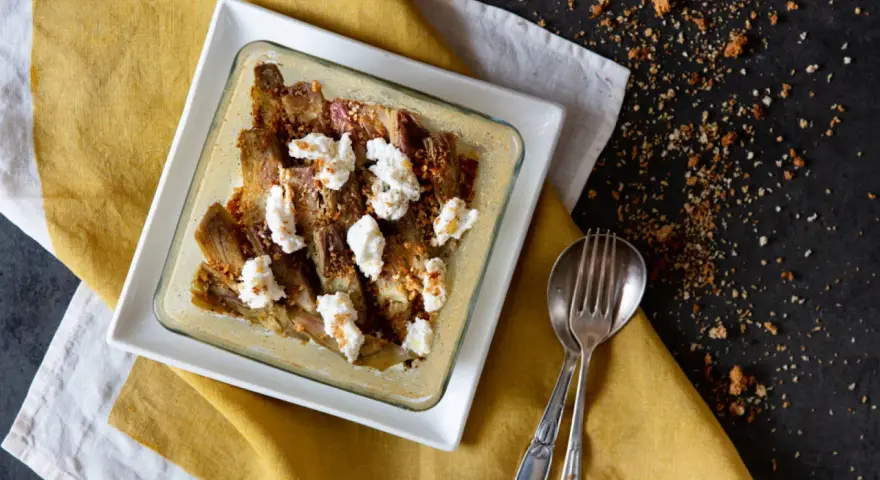 Carciofi e ricotta al forno in una teglia bianca, guarniti con pangrattato croccante. Serviti su un tovagliolo giallo.