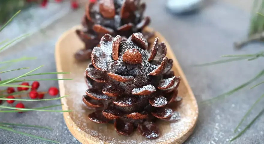 Deliziose pigne dolci decorate con zucchero a velo, su un vassoio di legno, con rami di pino e bacche rosse sullo sfondo.